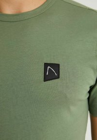 Olivegrünes T-Shirt aus weichem Stoff mit Rundhalsausschnitt, das mit einem schwarzen rechteckigen Patch mit einem weißen geometrischen Design versehen ist.