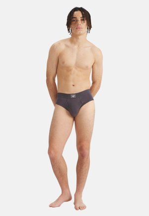 Junger Mann mit mittellangen Dreadlocks, der dunkelgraue Unterhosen trägt und barfuß vor weißem Hintergrund steht