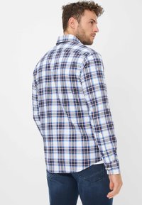 Uomo in camicia a maniche lunghe a quadri blu, bianca e nera, in piedi con la schiena rivolta, abbinata a jeans blu scuri davanti a uno sfondo semplice.