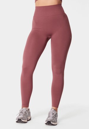 Kobieta ubrana w purpurowe legginsy z wysokim stanem i szare buty sportowe, stojąca na jednolitym białym tle.
