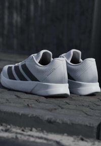 Paire de baskets blanches et grises avec l’étiquette "DURAMO RC 2", posée sur un pavé en pierre contre un fond texturé sombre.