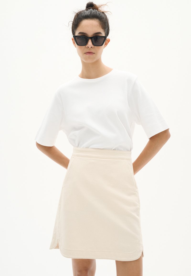 T-shirt en coton blanc à manches courtes associé à une jupe évasée beige avec fentes latérales et coutures structurées. Des lunettes de soleil noires complètent le look.