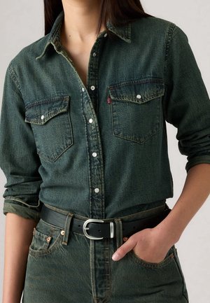Personne portant une chemise en denim vert foncé avec des poches poitrine et une ceinture noire, une main dans la poche du pantalon en denim assorti.