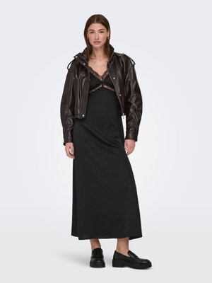 Donna che indossa un vestito a sottoveste nero con bordi in pizzo, una giacca corta in pelle marrone scuro e mocassini neri con plateau, in piedi su sfondo bianco.