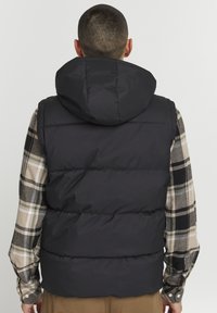 Padded black vest s kapucňou, so prešívaným dizajnom. Nosený cez kockovanú košeľu v béžovej, čiernej a zelenej farbe.