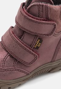 Lilla lærstøvle med to Velcro-remmer, teksturert overflate og en brun gummisåle. Har en logomerke på siden.