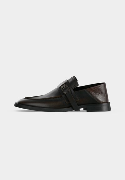 Mocassin en cuir marron foncé avec un bout arrondi, une bride et un détail de boucle, et une semelle noire basse sur fond gris.