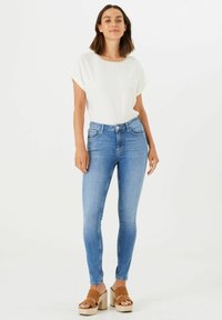 Jeans skinny azules de cintura alta, combinados con una blusa blanca de manga corta con un escote decorativo, y sandalias de plataforma marrones.