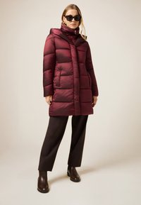 Burgundy dunjakke med hætte, med lynlåslukning, to forlommer og quiltet design. Båret over mørke bukser og støvler.