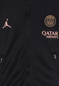 La chaqueta negra con cremallera presenta acentos en rosa, incluye el logo de Jordan y la insignia del PSG. La tela es suave con una ligera textura, y cuenta con bolsillos con cremallera.