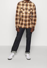 Uomo che indossa una camicia a quadri arancione, blu navy e bianca con bottoni, jeans scuri arrotolati, calzini bianchi e sneakers bianche con il baffo nero.