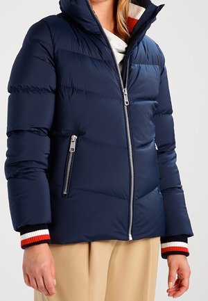 Veste matelassée bleu marine avec fermeture éclair argentée, poches zippées, col montant, portée sur une chemise blanche aux poignets rayés et un pantalon beige.