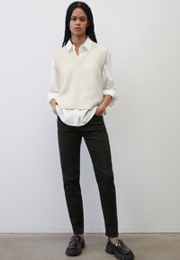 Off-white gestrickte Weste mit V-Ausschnitt über einem weißen Hemd mit Knöpfen, kombiniert mit schwarzen Slim-Pants und schwarzen Slippern mit dicker Sohle.