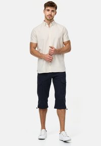 Light beige short-sleeve button-up shirt paired with knee-length navy cargo shorts. White sneakers complete the look. Cargo shorts feature pockets.