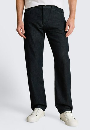 Mężczyzna w ciemnych jeansach o prostych nogawkach i białych trampkach, stoi z rękami swobodnie opuszczonymi wzdłuż ciała.