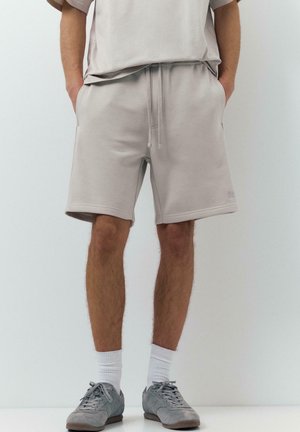 Uomo che indossa pantaloncini beige chiaro con coulisse, abbinati a calzini bianchi e sneakers casual grigie, in piedi contro uno sfondo bianco neutro.