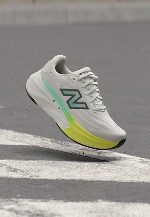 Λευκό και πράσινο αθλητικό παπούτσι New Balance στον αέρα πάνω από έναν γκρι δρόμο με λευκές διαβάσεις.