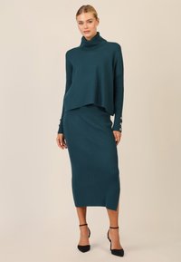 Maglione a collo alto lavorato a maglia color verde acqua, indossato su una gonna midi a coste verde acqua con spacco laterale, accentuato da dettagli con bottoni sulle maniche. Tacchi neri.