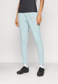 Light blue athletic pants with a slim fit, featuring a pink Nike logo on the left hip and a textured, breathable fabric. Black soccer cleats.