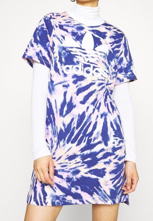 Osoba nosząca niebiesko-różową sukienkę Adidas z efektem tie-dye nałożoną na biały, długi rękaw golf, ręce swobodnie przy bokach.