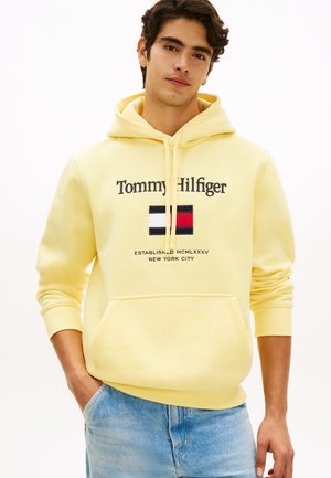 Jonge man die een gele Tommy Hilfiger hoodie en lichtblauwe spijkerbroek draagt, staand met één hand in de zak tegen een witte achtergrond.