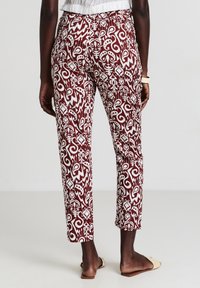 Donna che indossa pantaloni alla caviglia a strisce rosse e bianche, una maglietta bianca, sandali crema e un braccialetto chiaro, in piedi di spalle.