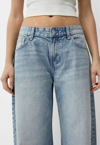 Jeans din denim de un albastru deschis, cu talie înaltă, picioare largi și buzunare frontale. Dispune de un închidere cu nasture și fermoar, cu o ușoară decolorare.