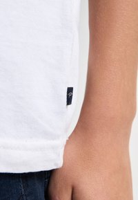 Hvid bomulds T-shirt med en syet kanten og et navy mærke med et guld bogstav "P" på venstre side nær bunden af kanten.