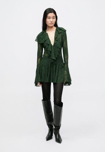 Πράσινο, διάφανο φόρεμα με βολάν, βαθύ V λαιμόκοψη και μακριά μανίκια. Συνδυασμένο με μαύρο εφαρμοστό κολάν και ψηλές, γυαλιστερές, μαύρες μπότες.