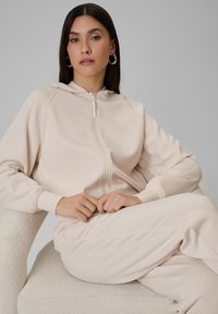 Femme aux longs cheveux foncés portant un sweat à capuche beige zippé et un pantalon assorti, assise sur une chaise texturée de couleur claire sur fond gris uni.