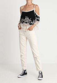 Haut noir décolleté épaules avec broderie florale blanche et détail à franges, associé à un pantalon beige clair droit et des baskets noires.