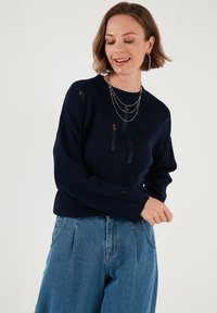 Tengerészkék kötött pulóver, megviselt részletekkel, hosszú ujjakkal és kerek nyakkivágással, magas derekú kék farmernadrággal párosítva.