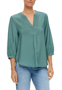 Turquoise blouse met een V-hals, driekwartmouwen en geplooide manchetten. Gemaakt van lichte, gestructureerde stof. Gemaakt in combinatie met blauwe jeans.