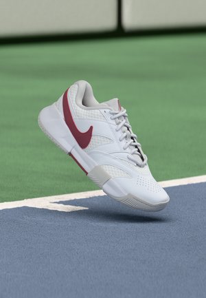 Witte en rode sportschoen zwevend boven het oppervlak van een tennisbaan nabij de witte begrenzingslijn.
