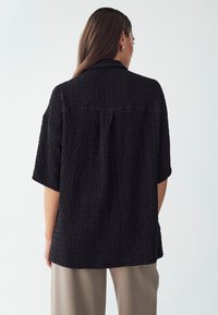 Camisa negra texturizada de espalda descubierta con manga corta y collar, que presenta un corte relajado y ligeras pinzas. Combinada con pantalones de tono neutro.