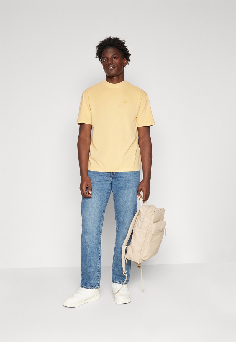 Żółta bawełniana koszulka z krótkimi rękawami, luźny krój. Zestawiona z niebieskimi dżinsami prostymi i beżowym plecakiem oraz białymi sneakersami.