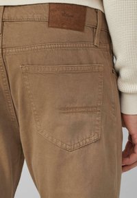 Nahaufnahme der hinteren Tasche einer Person mit braunen Hosen, auf der ein kleines Leder-Brandlabel zu sehen ist, sowie ein Teil des Ärmels eines cremefarbenen Strickpullis.