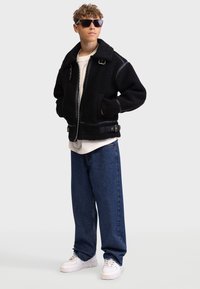 Schwarze Fleece-Bomberjacke mit einem Reißverschluss, mit Kragen, Seitentaschen und verstellbaren Riemen. Getragen über einem weißen Hemd und blauen Jeans.