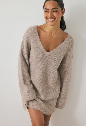 Knit sweater in light beige with a deep V-neck, oversized sleeves, and ribbed texture. Soft, fuzzy material with a relaxed fit.
