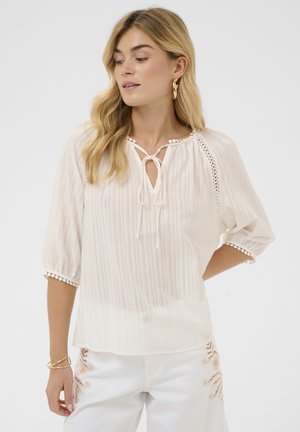 Szőke nő, laza hullámokkal, krémszínű blúzban, nyakkendős nyakkivágással és háromnegyedes ujjakkal, fehér nadrággal, mely virágos hímzéssel díszített.