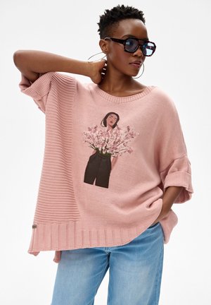 Vrouw draagt een oversized roze trui met een bloemmotief, grote ronde oorbellen, zonnebril en lichtblauwe spijkerbroek, poseert met hand achter haar nek.