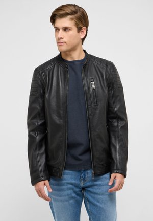 Junger Mann trägt eine schwarze Lederjacke, ein dunkelblaues Hemd und blaue Jeans und blickt leicht nach links vor schlichtem Hintergrund.