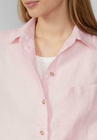 Chemise en lin rose pâle avec fermeture à boutons, col pointu, et une poche poitrine unique. Tissu texturé avec un léger éclat.