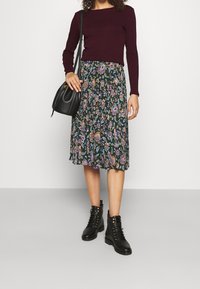 Bordeauxfarbener langärmeliger Oberteil kombiniert mit einem knielangen Rock im floralen Muster in dunklem Grün, getragen mit schwarzen Schnürstiefeletten und einer schwarzen Tasche.