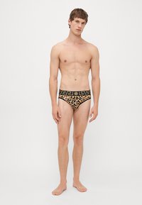 Junger Mann steht barfuß und trägt Leopardenmuster-Unterhosen mit einem schwarz-goldenen Musterbund vor einem weißen Hintergrund.