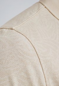 Tissu tricoté beige avec un motif texturé subtil, présentant une finition lisse et des détails de couture visibles.