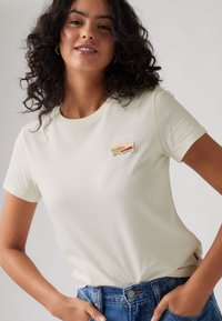Kortärmad krämfärgad t-shirt med rund hals, prydd med en färgglad broderad logotyp på bröstet. Tillsammans med högmidjade blå denimjeans.