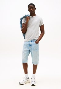 Ung mand iført grå t-shirt, lyseblå denimshorts, hvide sneakers, holder en denimjakke over skulderen, stående mod en hvid baggrund.