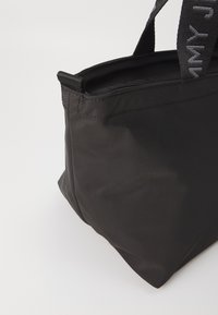 Schwarze Tragetasche aus glattem Material, mit einem Reißverschluss oben und doppelten Griffen, die Textakzente am Riemen aufweisen. Kompakte und strukturierte Form.