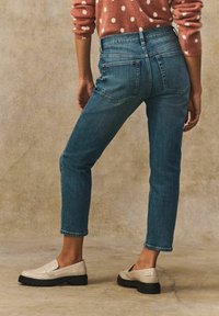 Vaaleansiniset farkut, joissa on kapea vyötärö, kapenevat lahkeet ja huomaamattomat takataskut; yhdistetty vaaleisiin, teksturoituihin loafereihin tasaisella pinnalla.